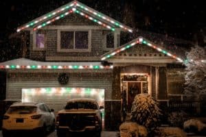 Snowy suburban house decorated with red, green, and white Christmas lights outlining the roof and garage. Two cars are parked in the driveway. How long do Christmas lights last? This house looks festive!