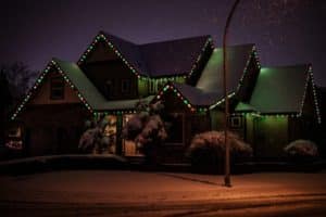 Snow-covered house adorned with red and green LED Christmas lights along the roofline. Tire tracks mark the snowy yard, illuminated by a street lamp, showcasing a charming DIY LED Christmas light installation.