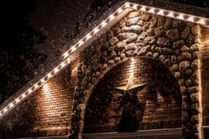 Snow falls on a brick and stone house in Naperville, IL, illuminated by warm holiday lights along the roofline. A large star decoration is centered above the entrance, highlighting a professional holiday light installation.