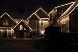 House with Christmas light installation outlining the roofline, set against a dark, snowy night. A vehicle is parked in the driveway, adding to the cozy, festive "Home" atmosphere.