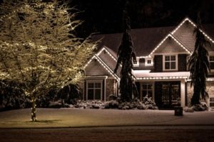 Warm white Christmas lights outline a snowy home and wrap a bare tree in the yard.