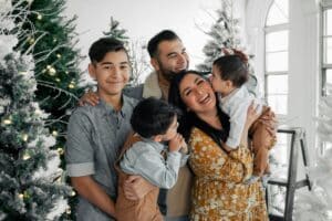 A happy family of five poses for a holiday portrait with Christmas trees, showcasing the joy of holiday guest preparation. The mother holds a baby who kisses her cheek, while another son hugs her leg.
