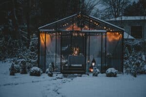 Snowy winter patio idea: A glass greenhouse glows with warm string lights, a cozy table set for a winter gathering amidst a snow-covered garden.