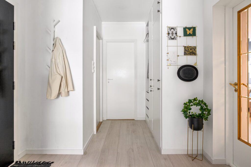 Bright, minimalist mudroom hallway. A light coat hangs on a white peg rack. A black hat sits above a potted plant on a gold stand. White storage cabinets line the hall, leading to a white door. Functional home mudroom design.