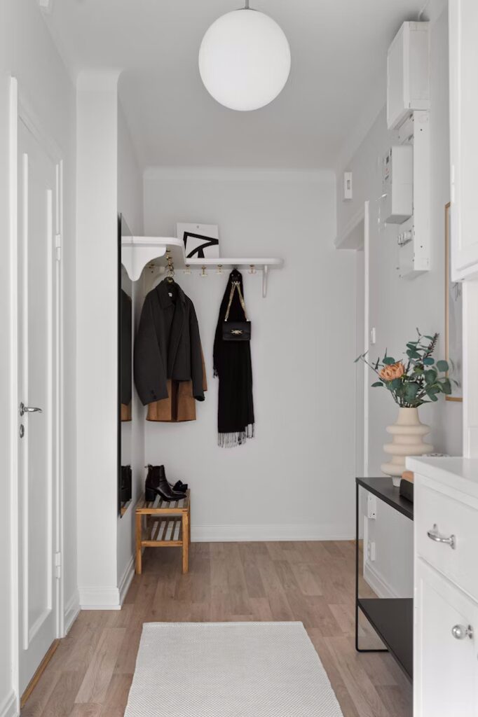 Bright, minimalist mudroom hallway with light wood floors, white walls, and a globe light. A coat rack holds a jacket, scarf, and handbag. A small stool and console table offer storage in this functional home mudroom.