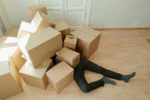 Person buried under a pile of moving boxes, only legs and feet visible. The scene humorously conveys the stress of moving and potentially needing professional help.