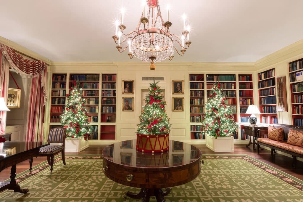 Elegantly decorated library for Christmas, featuring three lit Christmas trees adorned with red ribbons, bookshelves filled with books, a crystal chandelier, and a round wooden table on a patterned green rug.