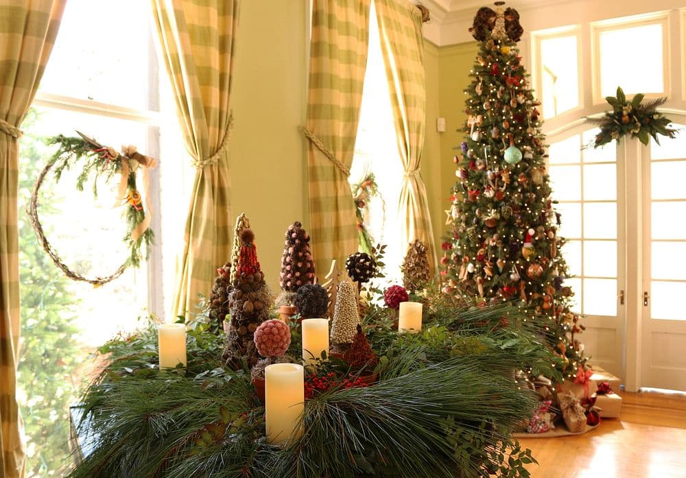 Festive Christmas decor featuring natural elements: pinecones, greenery, and candles arranged on a table in front of a window. A fully decorated Christmas tree stands nearby, embodying a cozy holiday ambiance.
