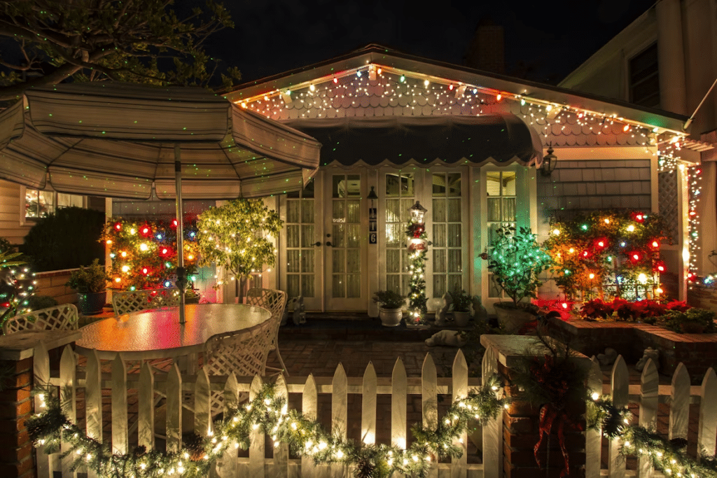 House exterior at night, beautifully decorated with Christmas lights outlining the roof and windows, and along the white picket fence. Outdoor patio furniture is illuminated by colorful Christmas lights on nearby bushes, showcasing how to hang Christmas lights.