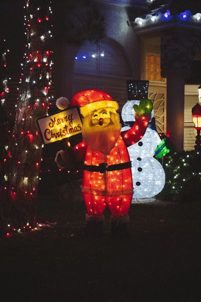 Festive Christmas light decoration: illuminated Santa holding a "Merry Christmas" sign stands next to a glowing snowman, with a light-wrapped tree and decorated house in the background.