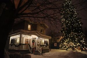 House decorated with Christmas lights, snow-covered yard, and a tall, brightly lit Christmas tree. Porch lights and garland add to the festive holiday scene.