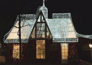 Santa's Workshop glows at night, illuminated with white Christmas lights outlining the roof and windows. Red decorations peek through the windows, creating a festive, warm ambiance.